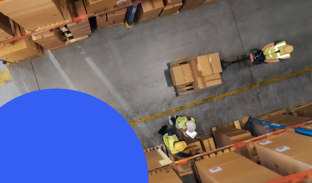 A birds-eye view of a warehouse aisle, with stacked boxes and people moving them around